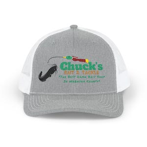 May include: A gray trucker hat with a white mesh back. The front features the text "Chuck's Bait & Tackle" in green and red, along with a black catfish illustration and fishing rod graphic. The text "The Best Damn Bait Shop In Wabasha County" is also included.