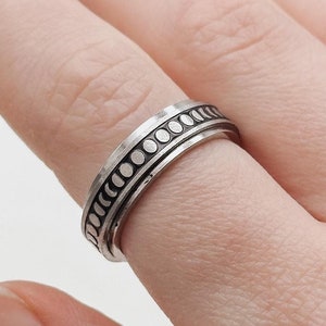 May include: A silver ring with a textured band and a row of small circles. The ring is worn on a finger.
