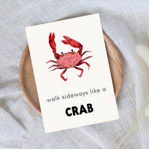 A 5x7 portrait flashcard with a trendy, bright red crab. The card reads, “walk sideways like a crab”. The card sits on top of a wooden bowl, which is on top of white linen.