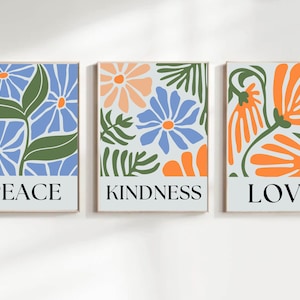 Three matching wall art prints featuring stylized floral designs and inspirational words. The left print showcases blue flowers and green leaves with the word 'PEACE' at the bottom. The center print displays a mix of blue, orange, and peach flowers and leaves with the word 'KINDNESS' at the bottom. The right print features orange flowers and green leaves with the word 'LOVE' at the bottom.  Each print has a light gray background.