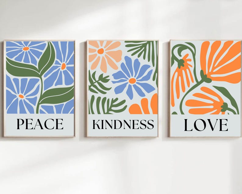 Three matching wall art prints featuring stylized floral designs and inspirational words. The left print showcases blue flowers and green leaves with the word 'PEACE' at the bottom. The center print displays a mix of blue, orange, and peach flowers and leaves with the word 'KINDNESS' at the bottom. The right print features orange flowers and green leaves with the word 'LOVE' at the bottom.  Each print has a light gray background.