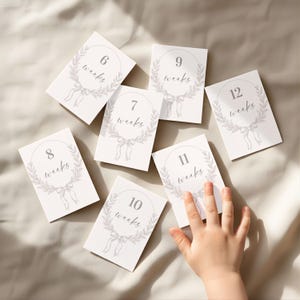 May include: White baby milestone cards with gray text and illustrations. Each card features the word "weeks" and a number from 6 to 12, surrounded by a wreath and bow design. A small child's hand is reaching for the cards.
