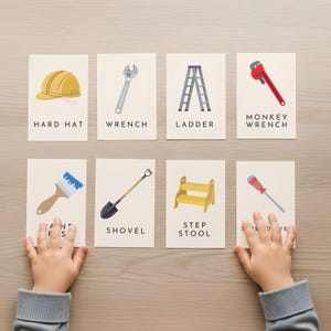 Puede incluir: Ocho tarjetas ilustradas con herramientas de construcción: casco, llave inglesa, escalera, llave de grifo, brocha, pala, taburete y destornillador. Cada tarjeta tiene el nombre de la herramienta. Las tarjetas están sobre una superficie de madera clara.