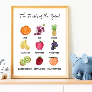 A framed print with the title 'The Fruits of the Spirit' and a list of nine fruits of the spirit with corresponding images. The fruits are love, joy, peace, patience, kindness, goodness, faithfulness, gentleness, and self-control. The images are of an orange, grapes, a pineapple, bananas, an apple, blueberries, kiwi, a strawberry, and a peach.