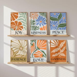 Six framed art prints with floral designs in orange, blue, and green hues. Each print features a word: Joy, Kindness, Peace, Patience, Love, and Goodness. The prints are displayed on wooden shelves against a white wall.