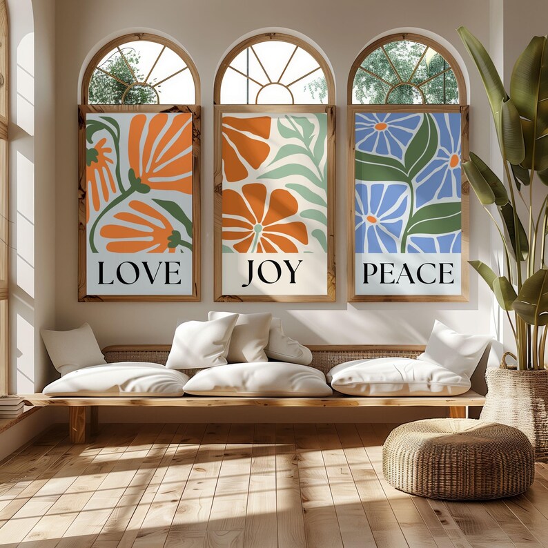 Bright and modern wall art trio of abstract floral prints reading “LOVE,” “JOY,” and “PEACE” in colorful mid-century style, displayed in sunlit arched window room.