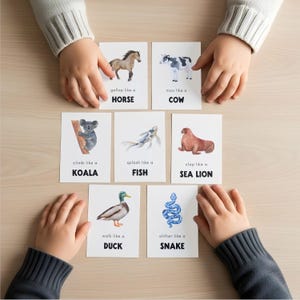 Educational flashcards featuring watercolor illustrations of animals. Each card displays an animal and a verb, such as "gallop like a HORSE" or "slither like a SNAKE." The cards are held by children's hands.