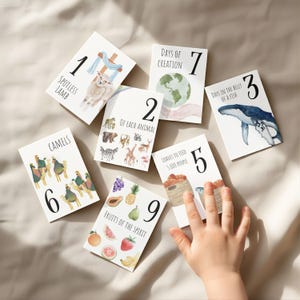 May include: A collection of illustrated cards with numbers and text. The cards feature images of a lamb, animals, a whale, camels, fruits, and more. The text includes "Spotless Lamb", "Days of Creation", and "Fruits of the Spirit". A child's hand is visible.