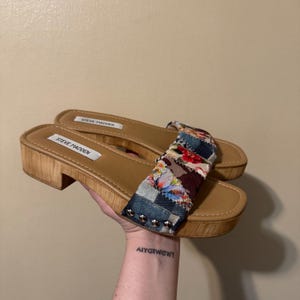May include: Steve Madden sandals with a wooden platform sole and a patchwork fabric strap. The strap features denim, floral, and patterned fabric. The sandals have a light brown footbed and are held in a hand.