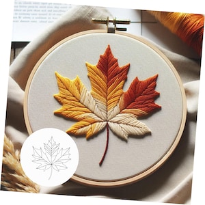 Peut inclure: Un gros plan d'un cerceau de broderie cousu à la main avec un motif de feuille d'automne coloré. La feuille est faite de fil orange, jaune et blanc et est entourée d'un cerceau en bois.