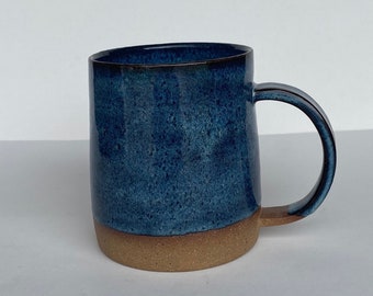 Könnte beinhalten: Eine blaue Keramiktasse mit braunem Rand und einer gesprenkelten Glasur. Die Tasse hat einen abgerundeten Körper und einen Henkel.