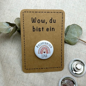 Könnte beinhalten: Eine braune Karte mit dem Text "Wow, du bist ein" und einem weißen Knopf mit einem Regenbogen und dem Text "Krippenkind Lina" darauf.