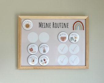 Könnte beinhalten: Eine weiße, gerahmte Magnettafel mit dem Text "Meine Routine" in Schwarz. Die Tafel hat ein Regenbogen-Design und mehrere bunte Knöpfe mit Bildern und Text in Deutsch. Die Knöpfe beinhalten "Guten Morgen", "Essen", "Hände Waschen", "Zähne Putzen", "Kindergarten", "Wickeln", "Anziehen" und Häkchen.