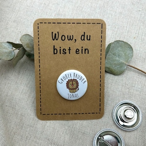 Könnte beinhalten: Ein weißer Button mit einer Cartoon-Löwen-Illustration und dem Namen "Jonas" ist an einer braunen Karte mit dem Text "Wow, du bist ein Großer Bruder" befestigt. Zwei zusätzliche Button-Rückseiten sind sichtbar.