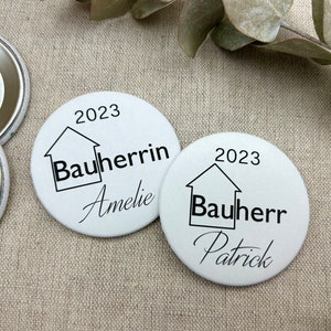 Könnte beinhalten: Zwei weiße runde Knöpfe mit schwarzem Text und einem Haussymbol. Der Text auf den Knöpfen lautet "2023 Bauherrin Amelie" und "2023 Bauherr Patrick".