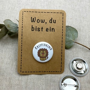 Könnte beinhalten: Eine braune Karte mit dem Text "Wow, du bist ein Krippenkind" und ein weißer Knopf mit einem Comic-Löwen, der eine Krone trägt. Der Knopf hat den Text "Krippenkind" am Rand.