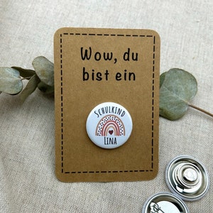 Könnte beinhalten: Eine braune Karte mit dem Text "Wow, du bist ein" und ein weißer Knopf mit einem Regenbogen und dem Text "Schulkind Lina".