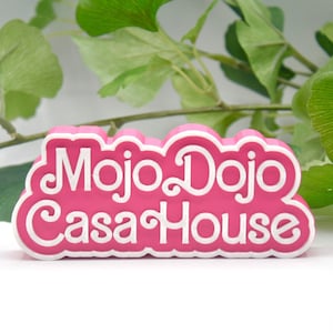 Puede incluir: Señal de plástico rosa con letras blancas que dice "Mojo Dojo Casa House".