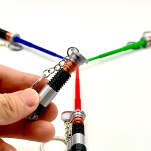 Puede incluir: Llaveros de sable de luz en miniatura en rojo, azul y verde. Los llaveros tienen mangos plateados y negros con un acento color cobre. Las hojas del sable de luz son de plástico y están unidas a un llavero plateado.