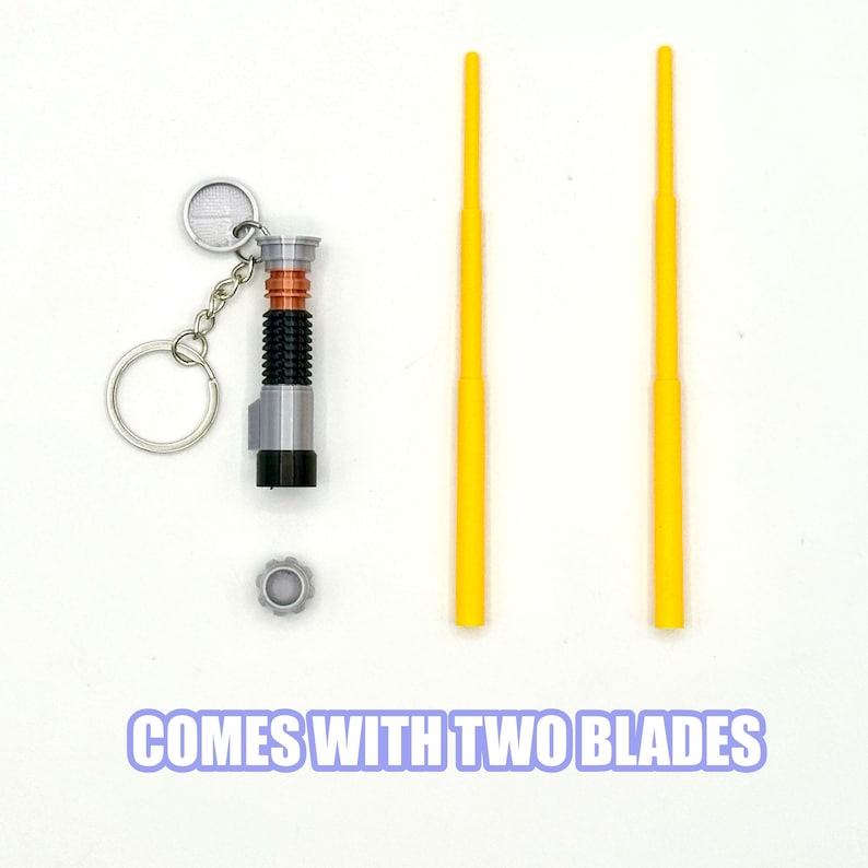 May include: A miniature lightsaber keychain with a silver finish, black grip, and orange accents. Two yellow blades and a small gray gear are also included. The text "COMES WITH TWO BLADES" is at the bottom.