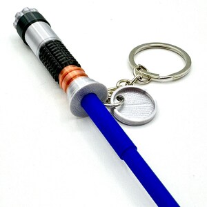 May include: A blue lightsaber keychain with silver, black, and copper accents. The keychain includes a silver keyring and a small, round tag. The lightsaber has a detailed handle and a bright blue blade.