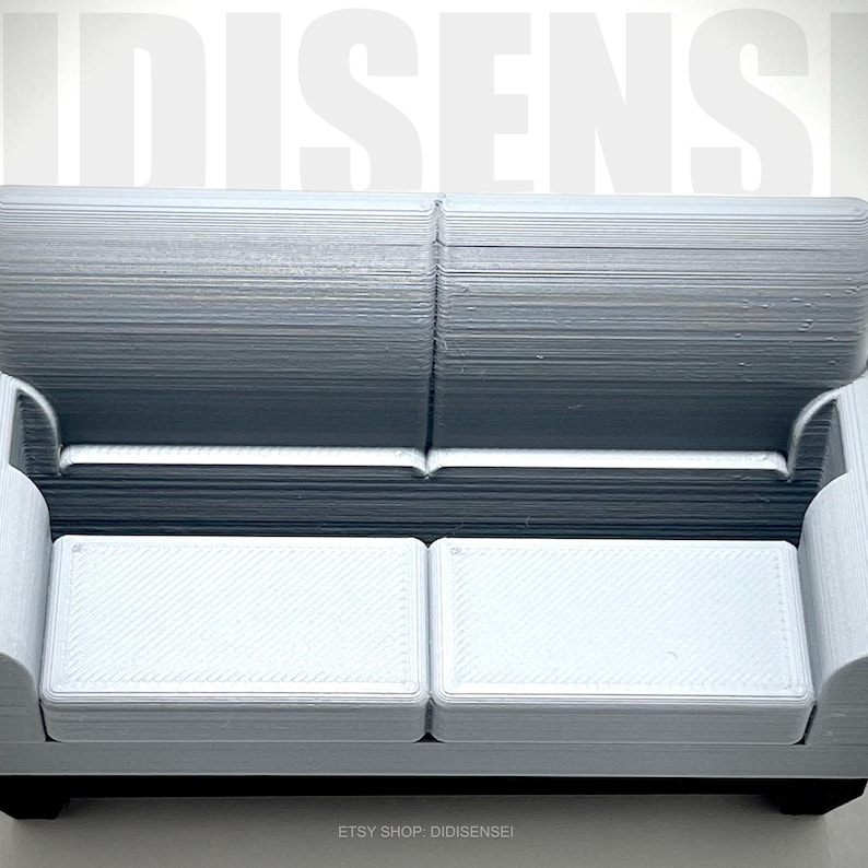 May include: A white miniature sofa with a textured fabric upholstery. The sofa has two cushions and a simple design.