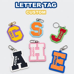 May include: Six custom letter tags featuring the names Grace, Sophia, Jacob, Aiden, Harper, and Ethan. Each tag is a different color combination with a metal lobster clasp or ring. The text LETTER TAG CUSTOM appears at the top of the image.