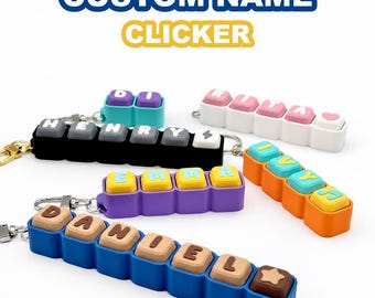 May include: Custom name clicker keychains featuring colorful rectangular bases with square, pressable buttons displaying letters and symbols. The set includes various color combinations like blue, purple, orange, black, and white with contrasting keys.