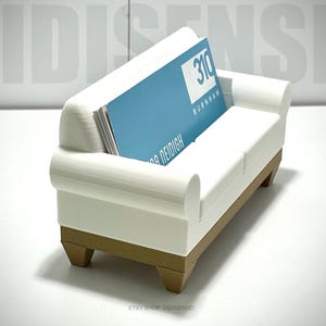 May include: White 3D printed couch shaped business card holder with a brown base. The holder has a slot for business cards and is holding a blue business card with the text "310 Burnham".