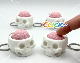 May include: Four white skull-shaped keychains with pink brain-shaped inserts. Each skull has hollow eye sockets and a nasal cavity. The pink brain inserts are textured and rounded. A metal keyring is attached to the side of each skull.