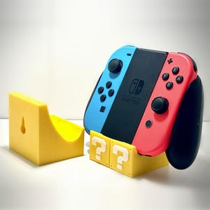 May include: A yellow 3D printed stand with two question mark blocks, holding a Nintendo Switch console with a red and blue Joy-Con controller.