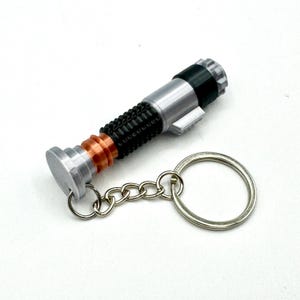 May include: A silver, black, and copper-colored lightsaber keychain. The keychain features a detailed replica of a lightsaber with a silver handle, black grip, and copper accents. A silver chain and keyring are attached.