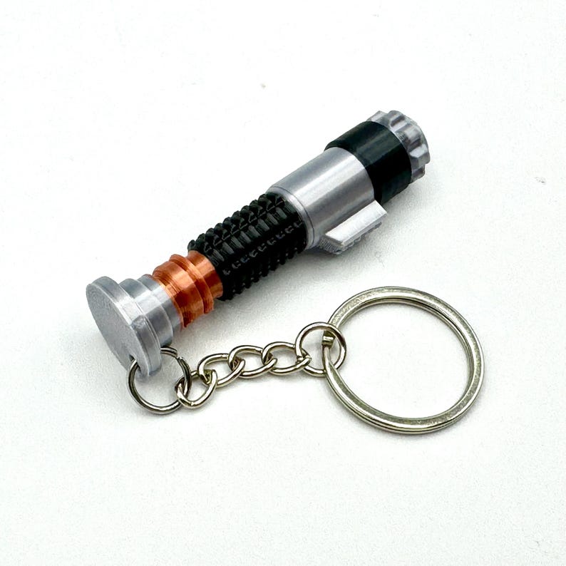 May include: A silver, black, and copper-colored lightsaber keychain. The keychain features a detailed replica of a lightsaber with a silver handle, black grip, and copper accents. A silver chain and keyring are attached.