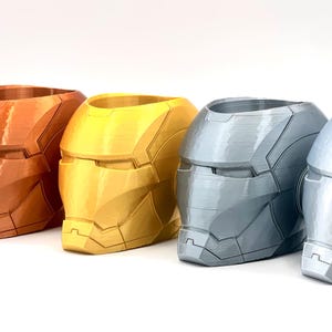 May include: Four 3D printed Iron Man helmet planters in different colors: copper, gold, silver, and black. The planters are made of plastic and have a smooth finish.