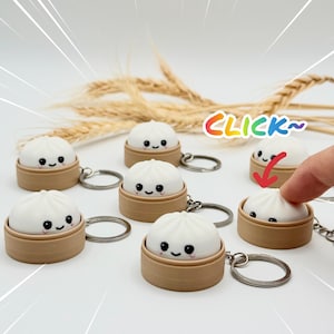 Peut inclure: Une collection de porte-clés en forme de raviolis blancs, chacun arborant un visage souriant et reposant dans un petit panier vapeur cylindrique beige. Les porte-clés incluent un anneau en métal. Le texte CLICK~ est visible en couleurs arc-en-ciel.