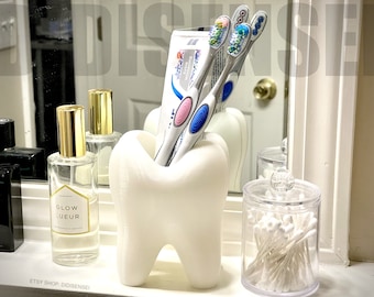 May include: White tooth-shaped toothbrush holder with three toothbrushes and a tube of toothpaste. The toothbrushes are blue and pink with white bristles. The toothpaste is white with blue and pink lettering. The holder is on a white countertop.