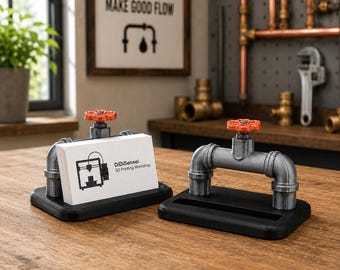 May include: Two business card holders designed to resemble industrial plumbing pipes. Each holder features a grey metallic pipe structure with a bright orange valve handle on top, mounted on a rectangular black base. One holds white cards.