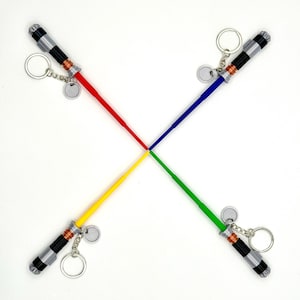 May include: Four lightsaber keychains arranged in a cross pattern. Each keychain has a different colored blade: red, blue, yellow, and green. The handles are silver and black with a small metal tag and keyring.