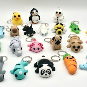 Peut inclure: Une collection de porte-clés colorés et faits à la main, représentant divers animaux et personnages. Les porte-clés comprennent un canard jaune, un pingouin noir et blanc, un bonhomme de neige, un carlin, un panda et une carotte. Chaque porte-clés a un anneau en métal argenté.