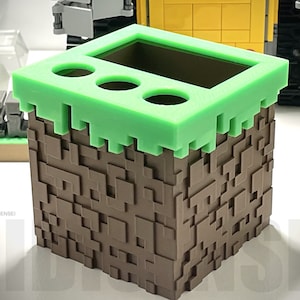 May include: A brown and green Minecraft-themed pencil holder with a grass top and three holes for pencils. The holder is made of plastic and has a pixelated design.