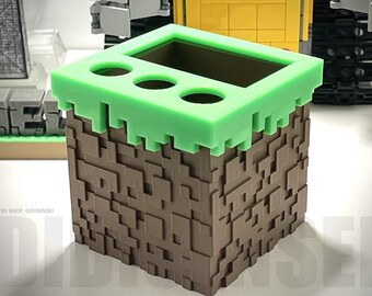 May include: A brown and green Minecraft-themed pencil holder with a grass top and three holes for pencils. The holder is made of plastic and has a pixelated design.