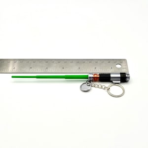 May include: A green lightsaber keychain with a silver and black handle, attached to a keyring. The lightsaber is approximately 10 inches long, with a green blade and a detailed hilt. A small, round silver charm hangs from the keychain.