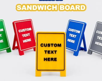 May include: A collection of five mini sandwich boards in green, red, yellow, blue, and gray. Each board features a rectangular frame with a stand and the text CUSTOM TEXT HERE printed on the front. The words MINI SANDWICH BOARD appear above.