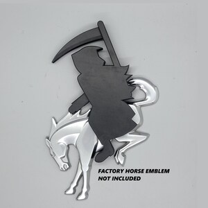 May include: A matte black silhouette of a grim reaper figure holding a scythe, positioned over a shiny chrome-finished horse emblem. The text FACTORY HORSE EMBLEM NOT INCLUDED is printed in black on the lower right side of the object.