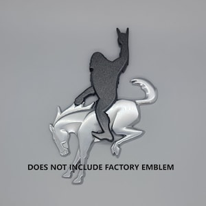 May include: A silver-colored emblem featuring a dark gray silhouette of Bigfoot riding a silver horse, with the hand gesture for rock and roll. The text "DOES NOT INCLUDE FACTORY EMBLEM" is printed below the emblem.