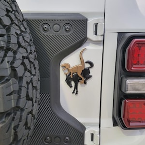 May include: A bronze-coloured dinosaur riding a black horse emblem is fixed to a white vehicle door. The emblem is mounted on a black textured panel. The vehicle's tyre and taillight are also visible.