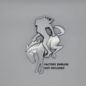 May include: Silver-colored metal emblem of a person riding a rearing horse. The emblem is a silhouette with a textured surface. The text "FACTORY EMBLEM NOT INCLUDED" is printed below the emblem.