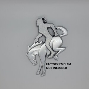 May include: Silver metal emblem of a person riding a rearing horse. The emblem is a silhouette of a person riding a horse. The text "FACTORY EMBLEM NOT INCLUDED" is below the emblem.