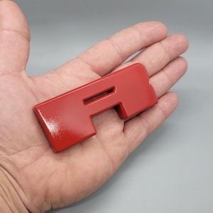 May include: A bright red, rectangular component with a central slot, held in a hand. The object has a smooth, glossy finish and an unusual shape, possibly a decorative piece or part of a larger assembly.