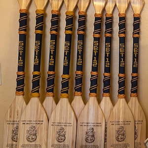 May include: A collection of wooden paddles with light brown blades and dark blue handles wrapped with gold and navy blue accents. Each paddle has the text "SET 12" and engraved text including "USN" and "September 27, 2024".